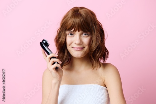 a happy, joyful, young woman stands with beautifully styled hair on a pink background and holds a black facial massager in her hand