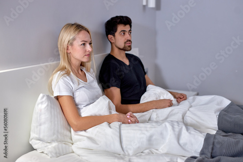 Couple watching tv in bed in cozy room at home looking at side, family concept. young caucasian man and woman in domestic clothes having rest, relaxing together. at weekends, holidays.