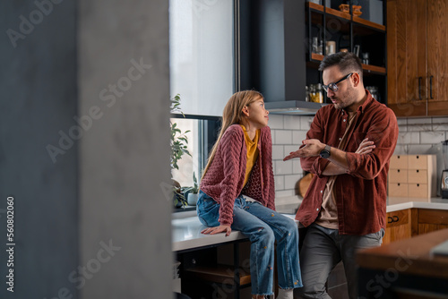 Young brown hair Caucasian father talking to his elementary age daughter while standing in the kitchen and gesturing with hands. Girl is sitting on a kitchen counter and carefully listening to him.