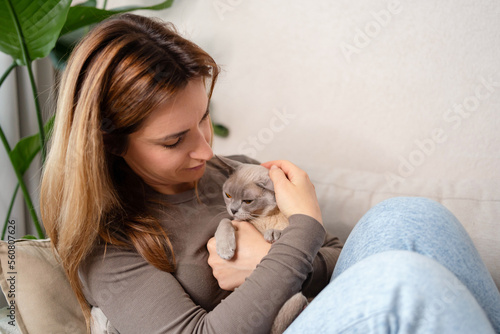 Beautiful cheerful young woman with a cute Burmese cat in her arms at home on the sofa, friendship love for pets.