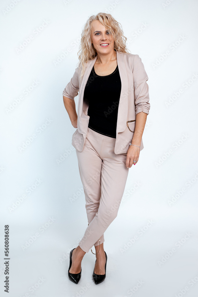 Full length photo of middle aged business woman with voluminous curly hairstyle in elegant beige pantsuit and black shoes, white background, copy space. Basic female clothes, casual office outfit Full length photo of middle aged business woman with voluminous curly hairstyle in elegant beige pantsuit and black shoes, white background, copy space. Basic female clothes, casual office outfit