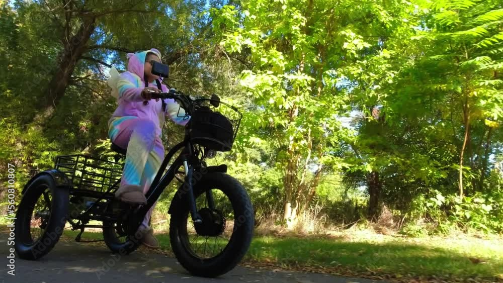 Woman in colourful candy rainbow bright unicorn pyjama or carnival dress riding an electric bicycle in the green park trail and having fun. Shiny sunny warm summertime day. Ecology concept. Woman in colourful candy rainbow bright unicorn pyjama or carnival dress riding an electric bicycle in the green park trail and having fun. Shiny sunny warm summertime day. Ecology concept.