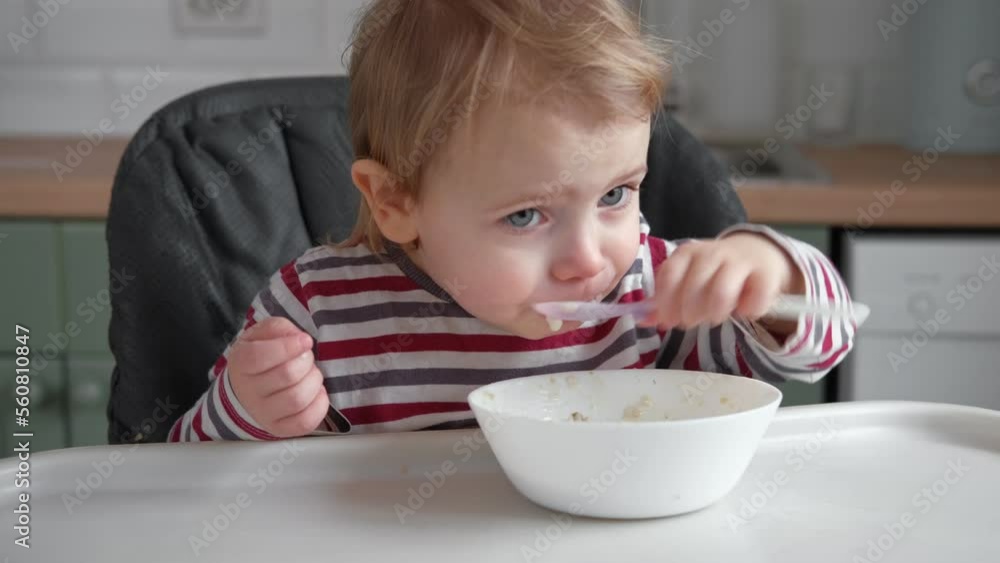 One year old hungry baby girl in striped casual clothes sits at white table in highchair and eats porridge herself with spoon. Blurred dining room background. Healthy eating for kids. Child nutrition