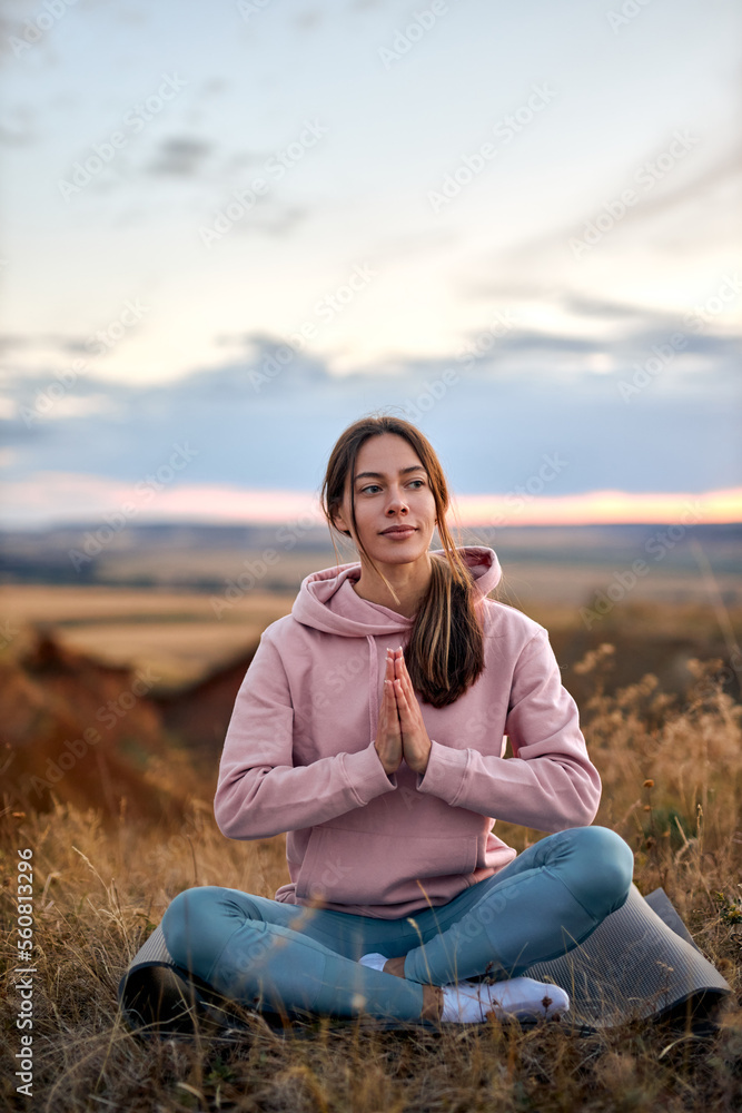 pretty caucasian female keep calm, meditating on fitness mat sitting outdoors in nature, keep hands together, engaged in yoga. young lady in sportswear concentrated on mind, enjoying yoga