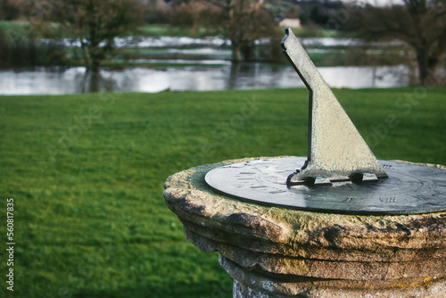 An old metal sundial set on a stone plinth or pedestal on a grassy lawn with a lake in the background