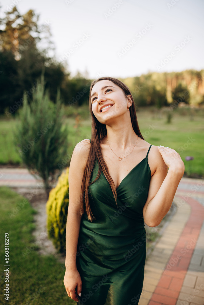 Beautiful smiling girl in a dark green dress with straps. Cute model girl stands on green grass and smiles. Brunette in the summer at a party. Emerald satin dress Beautiful smiling girl in a dark green dress with straps. Cute model girl stands on green grass and smiles. Brunette in the summer at a party. Emerald satin dress