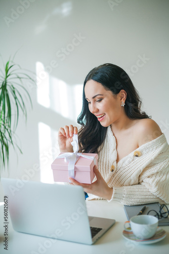 Young beautiful dark-haired woman of 34 years old in knitted sweater is talking on video link and holding pink gift box in her hands. Valentine's Day. Model received gift.