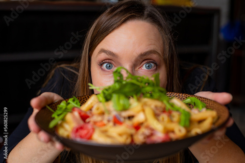 Portrait of middle-aged woman staring wide-eyed at camera, showing tasty pasta with cherry tomatoes, arugula and cheese.