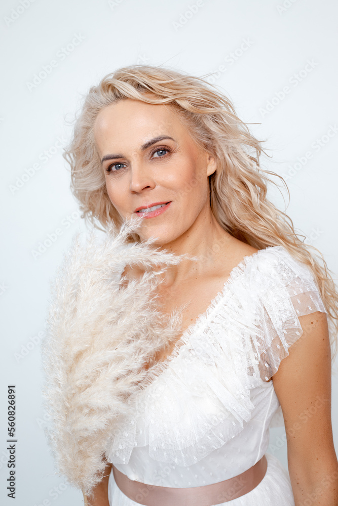 Smiling 50s mid aged adult woman with curly hairstyle and decorative feather in white dress isolated on white, copy space. Mild menopause, reproductive capacity, female wellness, lifting makeup Smiling 50s mid aged adult woman with curly hairstyle and decorative feather in white dress isolated on white, copy space. Mild menopause, reproductive capacity, female wellness, lifting makeup