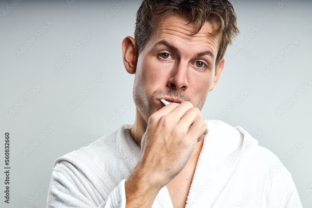 confused guy brushing tooth in early morning, after waking up. isolated on gray. caucasian man in white bathrobe look at camera with frustrated facial expression, having no enough sleep