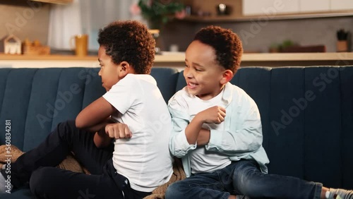 Two cute little african american boys brothers have conflict competition and turned away from each other with crossed arms sitting on sofa togetheк Adorable children kids argued at home 