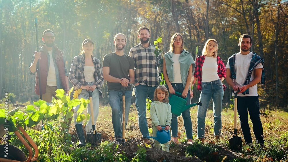 Portrait of Caucasian team of eco activists, men and women standing together in park with seedlings of trees and small kid and posing to camera. Outdoors. Males, females and child planting tree. - obrazy, fototapety, plakaty 