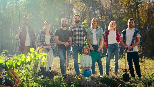 Portrait of Caucasian team of eco activists, men and women standing together in park with seedlings of trees and small kid and posing to camera. Outdoors. Males, females and child planting tree.