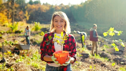 Portrait shot of blonde young beautiful Caucasian woman standing outdoor with tree seedling in pot and smiling cheerfully. Pretty female eco activist working against deforestation. Gardening. photo