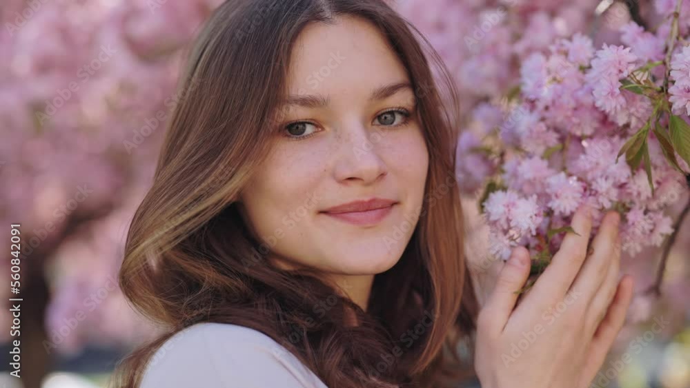 Portrait of beautiful caucasian young woman with natural brown hair looking at camera while touching tenderly blooming branches from sakura tree. Concept of femininity and beauty Portrait of beautiful caucasian young woman with natural brown hair looking at camera while touching tenderly blooming branches from sakura tree. Concept of femininity and beauty