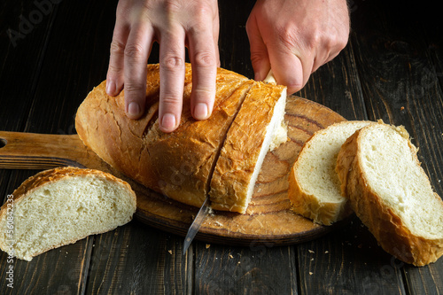 Cook's hand with a knife for cutting whole grain bread on a kitchen wooden board. Fresh bread on the table close-up. Wheat bread on the kitchen table or healthy food and traditional bakery concept