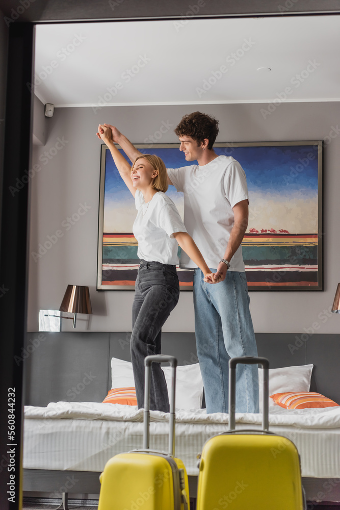 young and cheerful couple holding hands and having fun while standing on bed in modern hotel apartments.