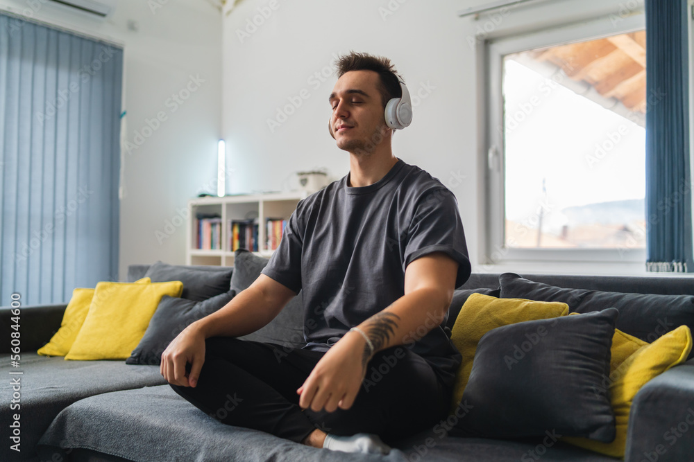A young guy is sitting on couch meditating and stretching while listening to music and using his mobile phone in his house during the day A young guy is sitting on couch meditating and stretching while listening to music and using his mobile phone in his house during the day