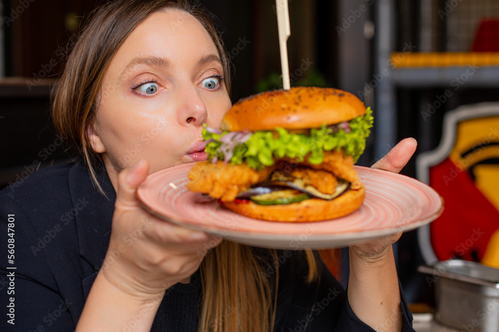 Portrait of surprised woman staring wide-eyed at burger with lettuce, chicken, onion on big plate in cafe restaurant.