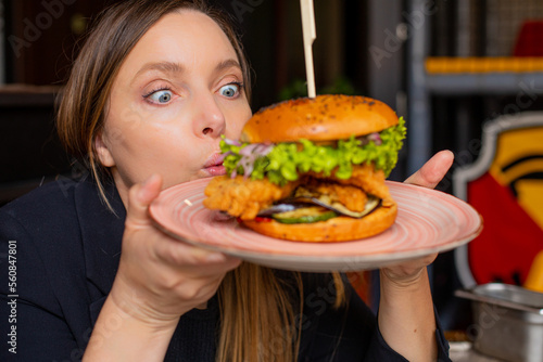 Portrait of surprised woman staring wide-eyed at burger with lettuce  chicken  onion on big plate in cafe restaurant.