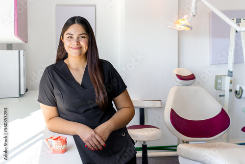 Mujer latina joven Dentista de pie recargada en consultorio Dental Sonriente  photo