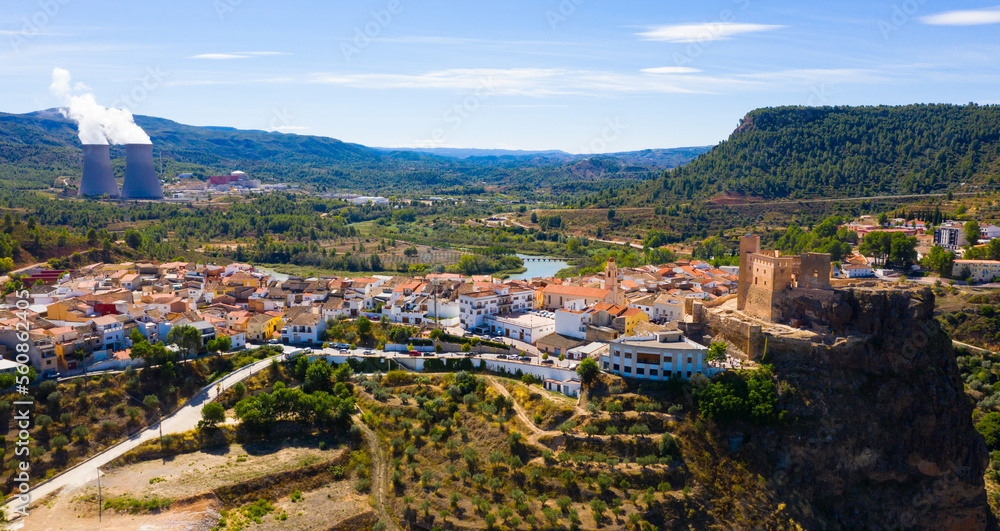 Aerial view of historical Spanish town of Cofrentes with medieval castle on top of basalt rock and modern nuclear power plant cooling towers on background, Valencia, Spain