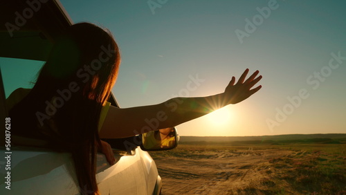 Free young woman travels by car catches wind with her hand from car window. Vacation. Girl with long hair is sitting in front seat of car, stretching her arm out window, catching glare of setting sun