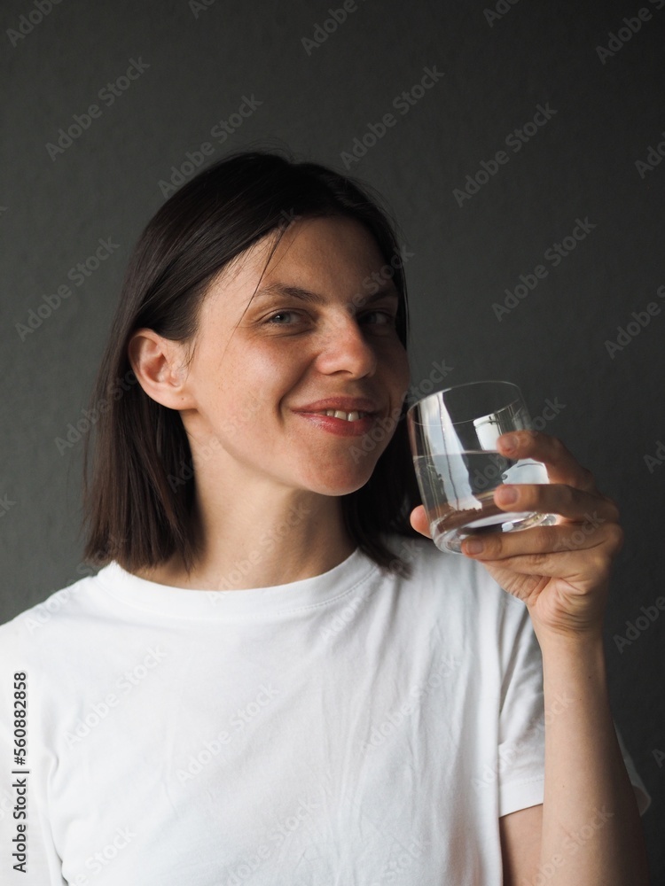 Young brunette woman wearing white t-shirt isolated over grey background drinking still water from glass and looking at camera,smilling.Closeup image of young woman holding a glass of water to drink. Young brunette woman wearing white t-shirt isolated over grey background drinking still water from glass and looking at camera,smilling.Closeup image of young woman holding a glass of water to drink.