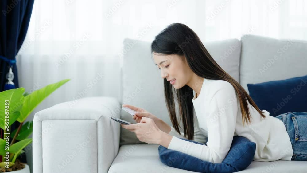 Young asian woman lying on sofa in living room, makes online banking payments through the internet from bank card on smartphone. Shopping online on mobile phone with credit card Young asian woman lying on sofa in living room, makes online banking payments through the internet from bank card on smartphone. Shopping online on mobile phone with credit card