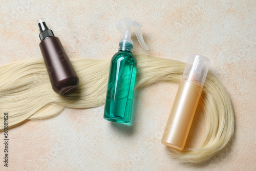 Spray bottles with thermal protection and lock of blonde hair on beige textured table, flat lay