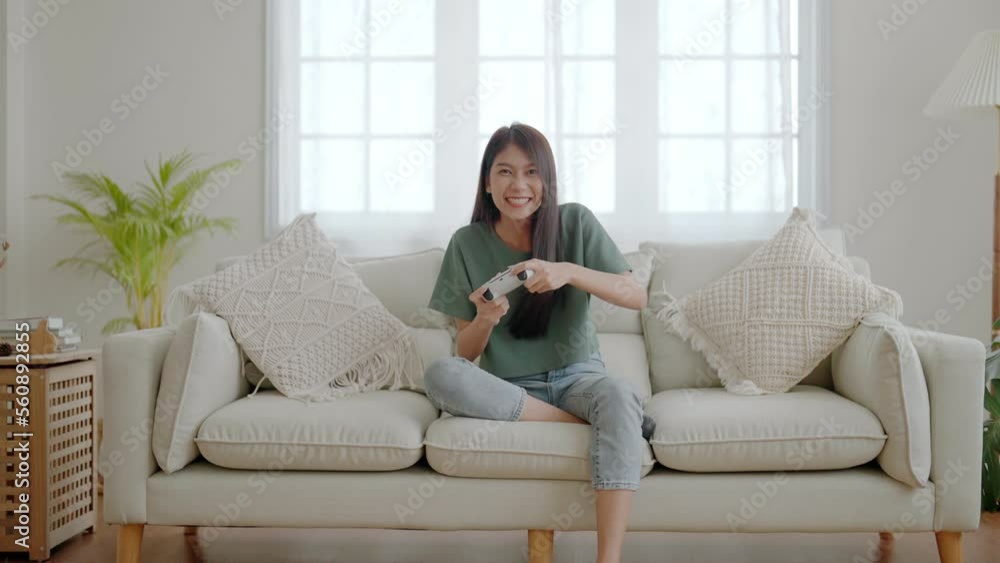 Beautiful young asian woman sitting on sofa and holding joystick playing online video game in living room at house Beautiful young asian woman sitting on sofa and holding joystick playing online video game in living room at house