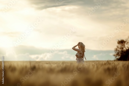 Profile portrait of blonde girl in golden dress, holding her hand to her head, standing in wheat field against of evening sky. Country style.
