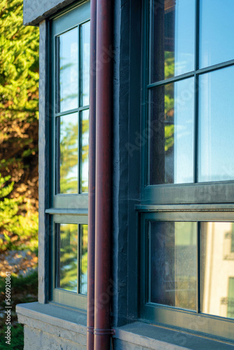 Visible windows on front of house or building with dark red metal rain gutter pipe and background sunny trees