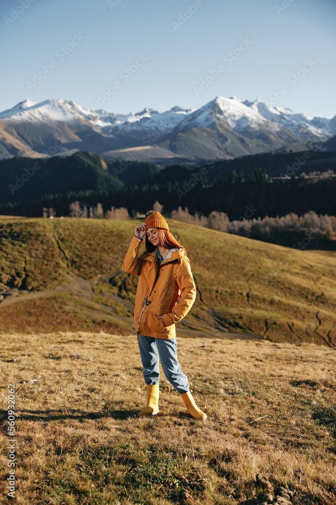 Autumn woman walking up the hill in full smile travels to the mountains in nature hiking and happiness in a yellow cape against the snowy mountains in the sunset, freedom of life style