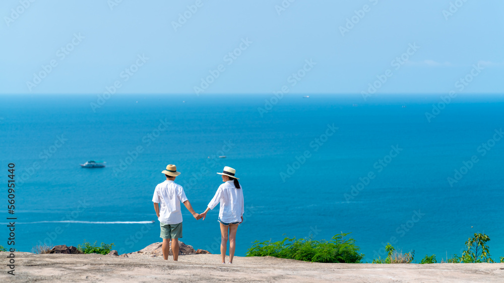 Happy Asian family couple enjoy outdoor lifestyle travel tropical island on summer holiday vacation. Husband and wife holding hands at mountain peak and looking nature and blue ocean in sunny day.
