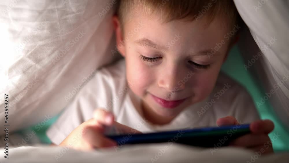 Portrait of face caucasian baby boy, playing in games on holding in hands mobile phone. Child lying in bed in room and enjoy process of game. Kid serious looking in screen and control with fingers. Portrait of face caucasian baby boy, playing in games on holding in hands mobile phone. Child lying in bed in room and enjoy process of game. Kid serious looking in screen and control with fingers.