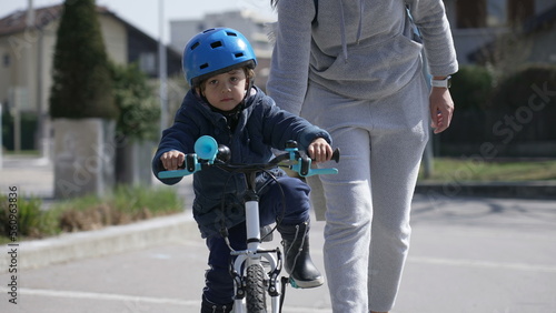 Child learning to ride bicycle outdoors with parent help. Little boy wearing helmet protection and coat rides bike during fall season