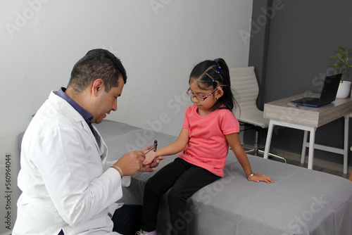 Fototapeta Naklejka Na Ścianę i Meble -  Latino doctor medic and girl patient in medical office checks her reflexes on hammer in her checkup to find disease diagnosis
