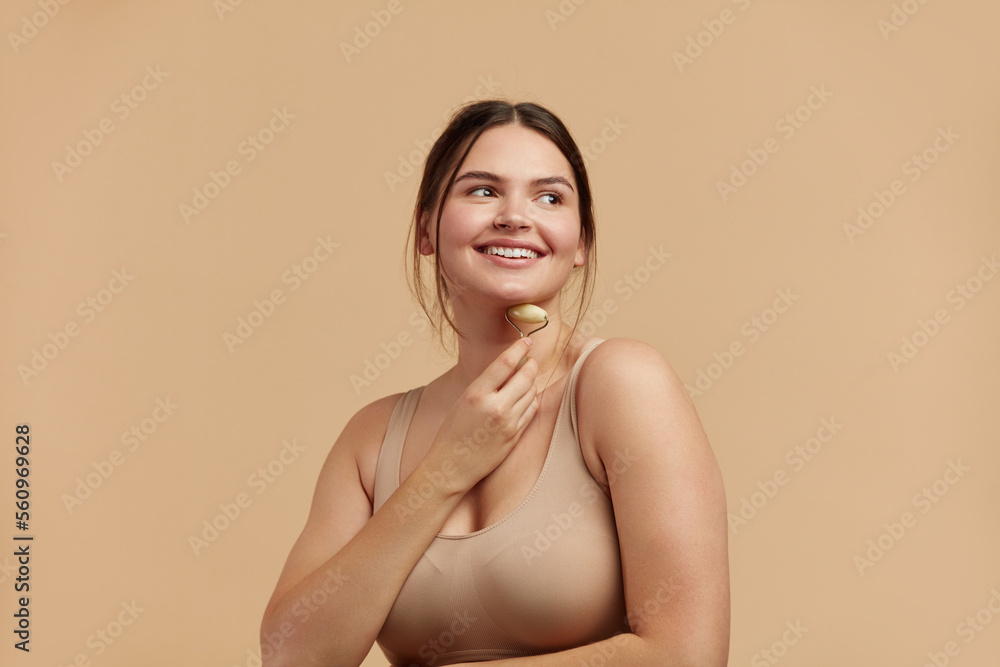 Obraz premium Face massage. Plus Size Woman Holding Jade Facial Roller for Skin Care, Beauty Treatment on Beige Isolate Background. Girl Taking Care of Skin with Natural Massager Closeup Portrait 