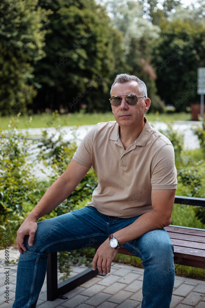 Obraz premium Vertical modern handsome relaxed confident mature gray haired businessman in sunglasses, shirt, wristwatch sit on bench
