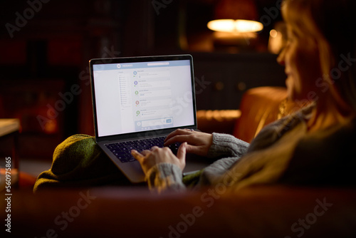 Woman At Home In Lounge Lying On Sofa With Cosy Fire Checking Emails On Laptop