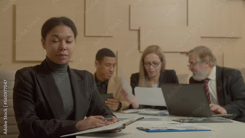 Portrait of successful charming black Asian female manager looking with radiant smile, expressing positivity and confidence over blurry diverse multiethnic business colleagues working in background.