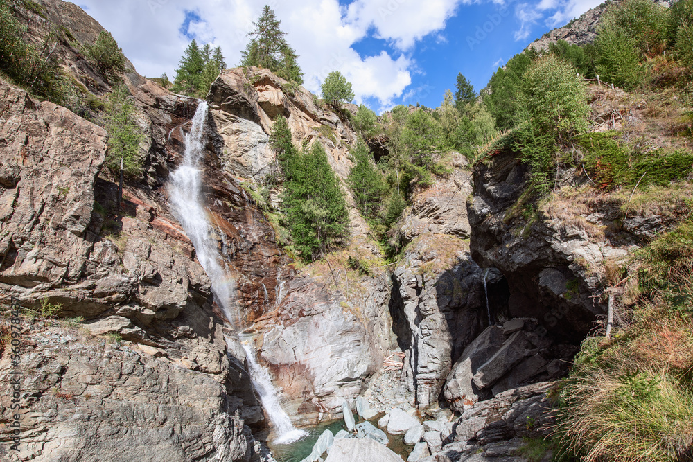 Obraz premium Bottom view of flowing Lillaz waterfall (Cascate di Lillaz) and granite karst rocks with evergreen trees under blue sky with white clouds, Aosta Valley, Italy
