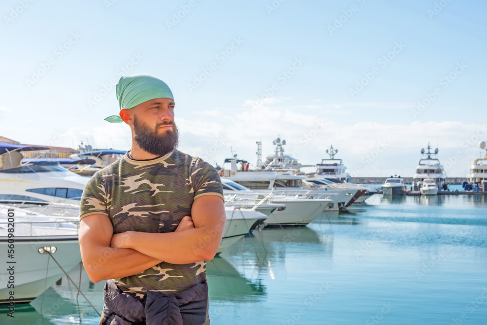 Obraz premium Handsome male beard turquoise bandanna military shirts stands hands folded on pier against the backdrop yachts in port bay in sunny weather.