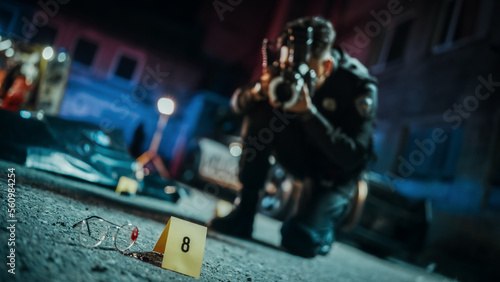 Policeman Taking Photos of Marked Evidence on a Crime Scene at Night. Forensics Police Officer Finds Glasses Potentially Belonging to the Dead Victim and Photographs it for Analysis. Focus on Glasses © Gorodenkoff