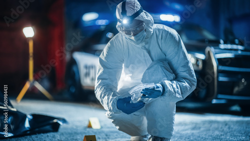 Portrait of Two Forensics Experts Doing Fieldwork at Night at a Crime Scene. One Technician Packs the Bloodied Knife as Murder Weapon While the Other is Taking Photos of the Dead Body.