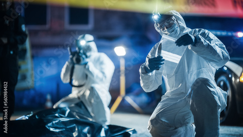 Portrait of Two Forensics Experts Doing Fieldwork at Night at a Crime Scene. One Technician Taking Photos of the Dead Body While the Other Packs the Bloodied Knife as Murder Weapon. photo
