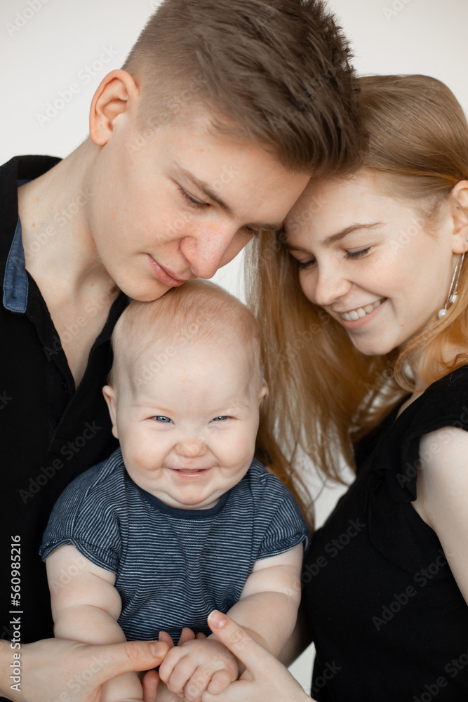 Portrait of happy family from three in black looks on white background. Mom and dad gently touch little hands of joyful smiling infant baby closeup. Parental affection, childcare, firstborn Portrait of happy family from three in black looks on white background. Mom and dad gently touch little hands of joyful smiling infant baby closeup. Parental affection, childcare, firstborn
