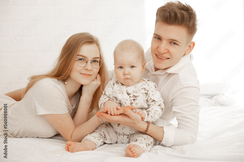 Happy young couple with little baby lie on bed and holding palms of each other, white background, free copy space. Home family photo of mom, dad and infant child. Parental affection and love Happy young couple with little baby lie on bed and holding palms of each other, white background, free copy space. Home family photo of mom, dad and infant child. Parental affection and love