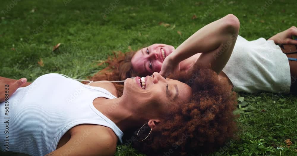 Portrait of Two Multiethnic Women Lying Down on a Park Lawn Using Earbuds to Listen to Music. Black Female Teenager Sharing her Favorite Song with her Friend While They are Sunbathing on a Hot Day Portrait of Two Multiethnic Women Lying Down on a Park Lawn Using Earbuds to Listen to Music. Black Female Teenager Sharing her Favorite Song with her Friend While They are Sunbathing on a Hot Day